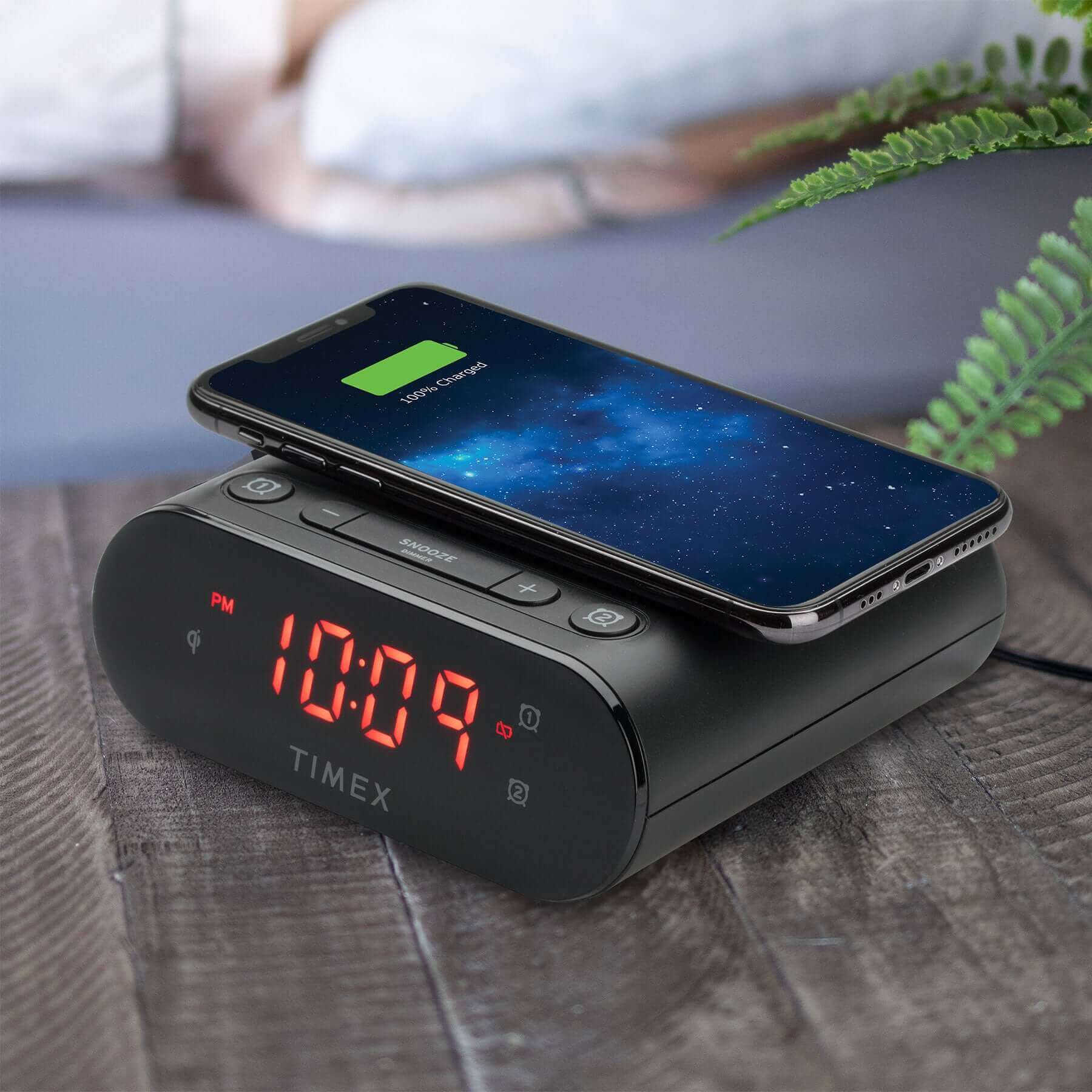 Timex TW300 Alarm Clock with Wireless Charging and Dual Alarms, shown charging a smartphone on a bedside table with dimmable display