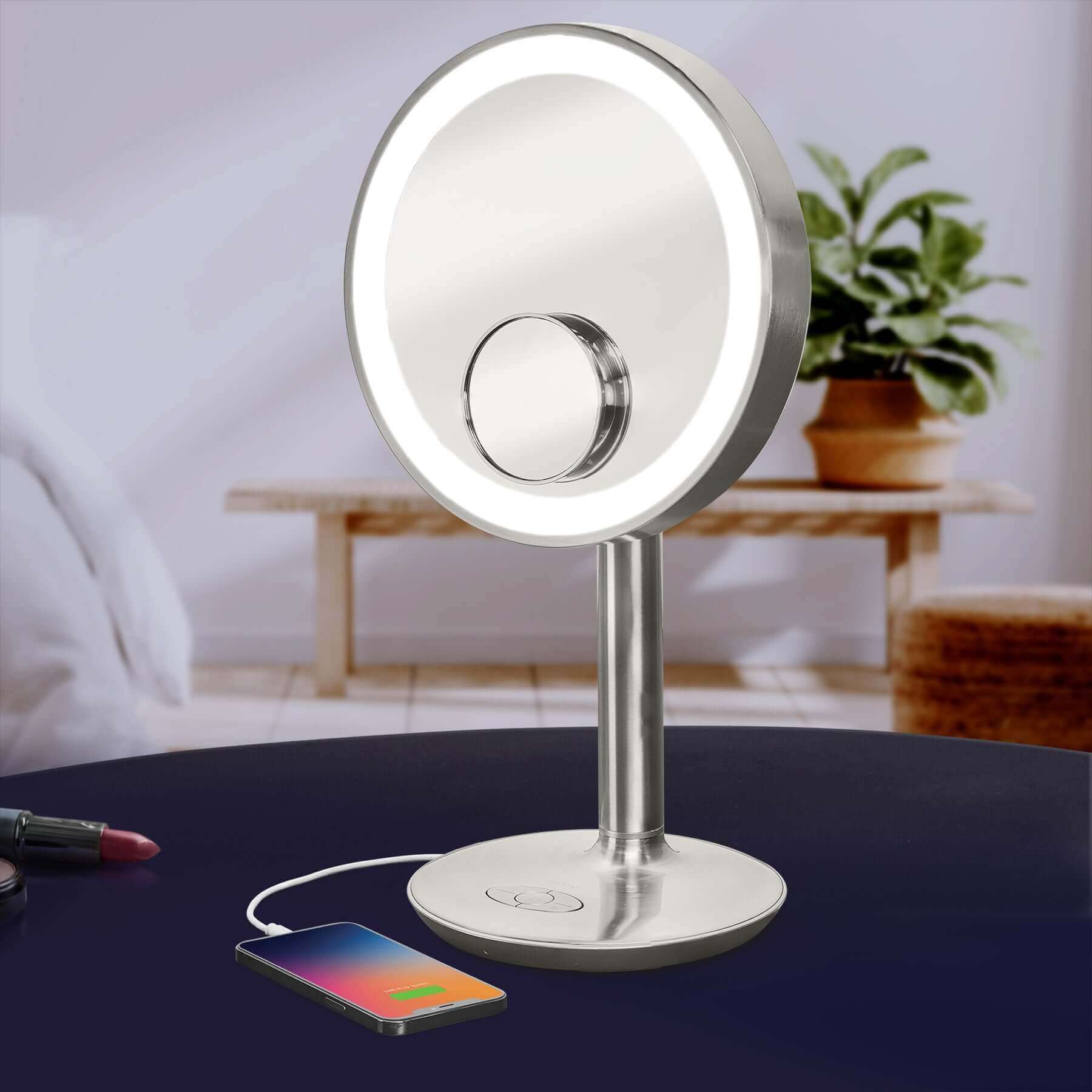 Vanity mirror with lights, wireless speaker, USB charging, and detachable magnifying mirror on table, next to a charging phone.