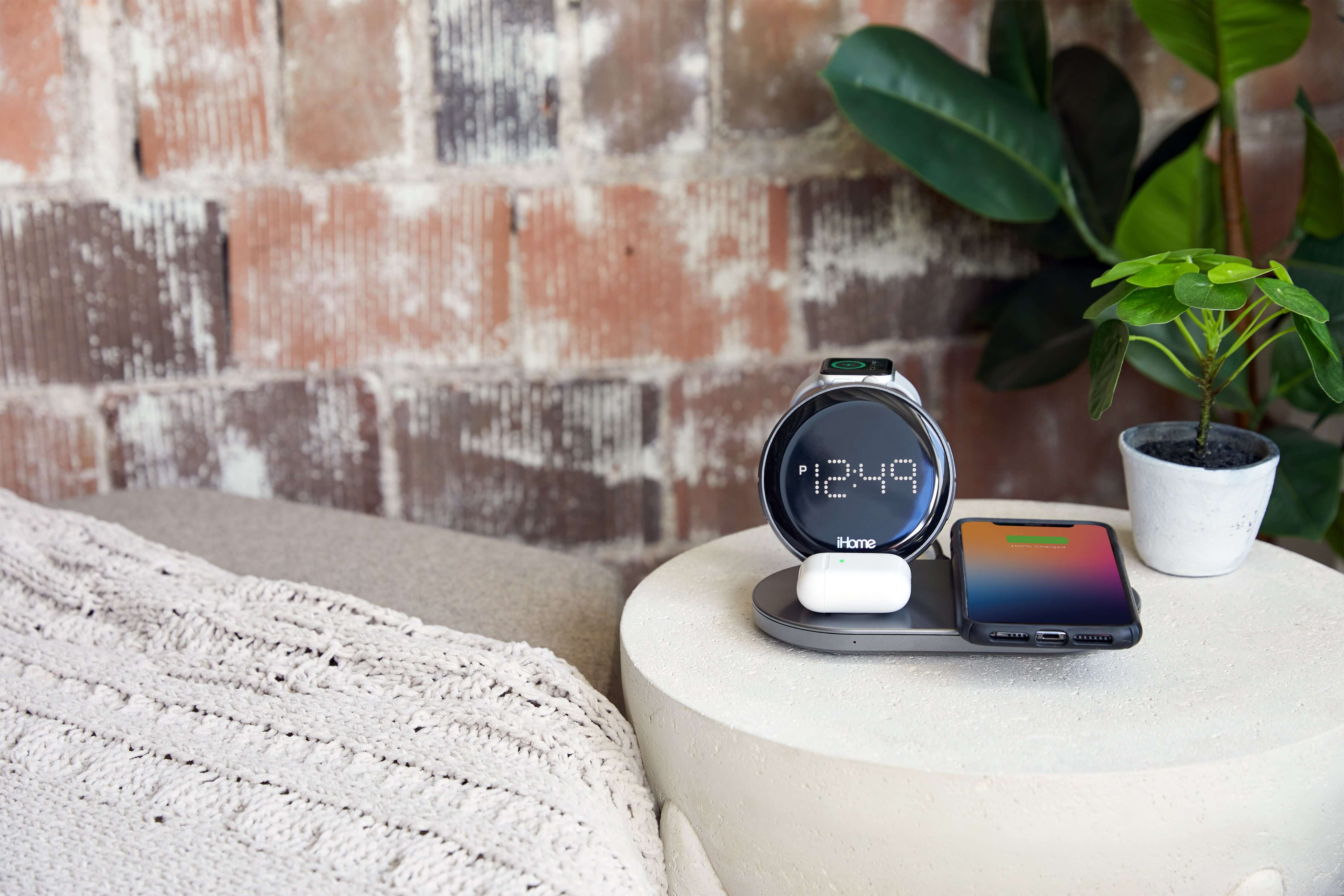 Smartwatch, smartphone, and earbuds charging on a sleek wireless charging pad on a side table with a cozy blanket and potted plant nearby.